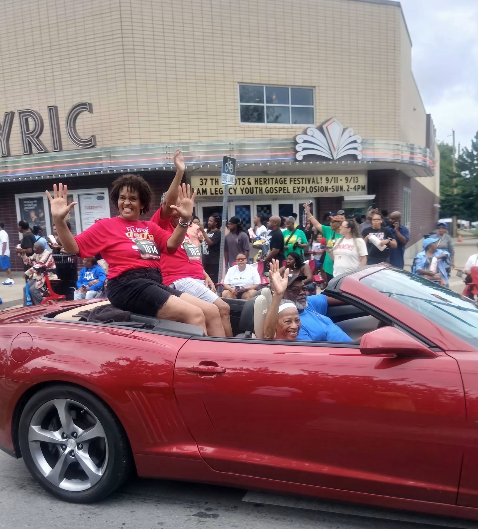 Parade participants riding in a red convertible and waving outside the Historic Lyric Theatre